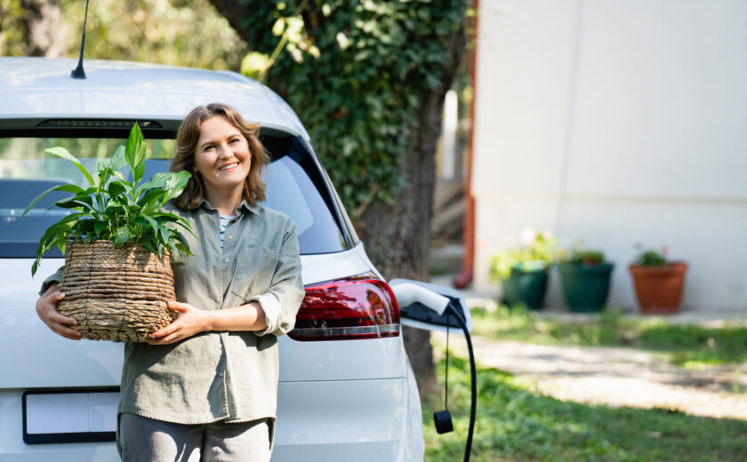 Woman with plant in pot next to a charging electric car in the yard of a country house